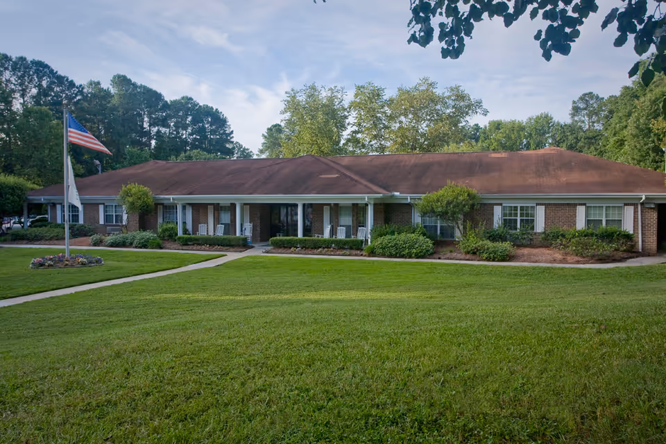Single-story brick building with a brown roof surrounded by green grass and trees. There is a flagpole with an American flag and another flag in front of the building. The building has several windows with white shutters and a covered porch with white rocking chairs.
