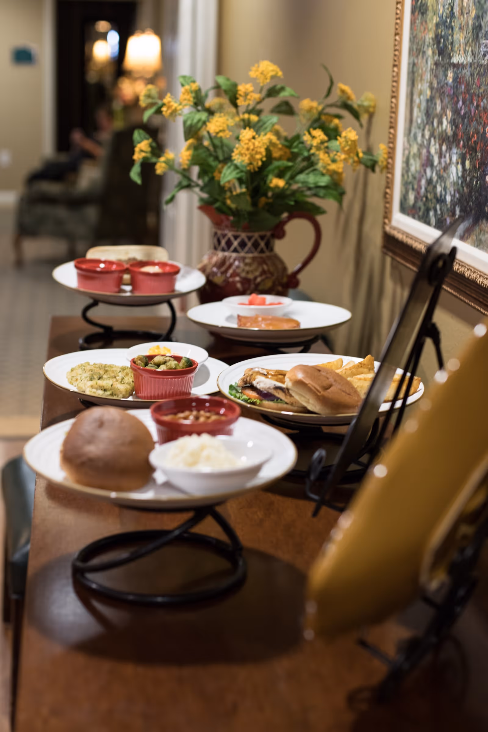 A variety of plated meals arranged on a wooden surface with a decorative vase of yellow flowers in the background and a framed painting on the wall.