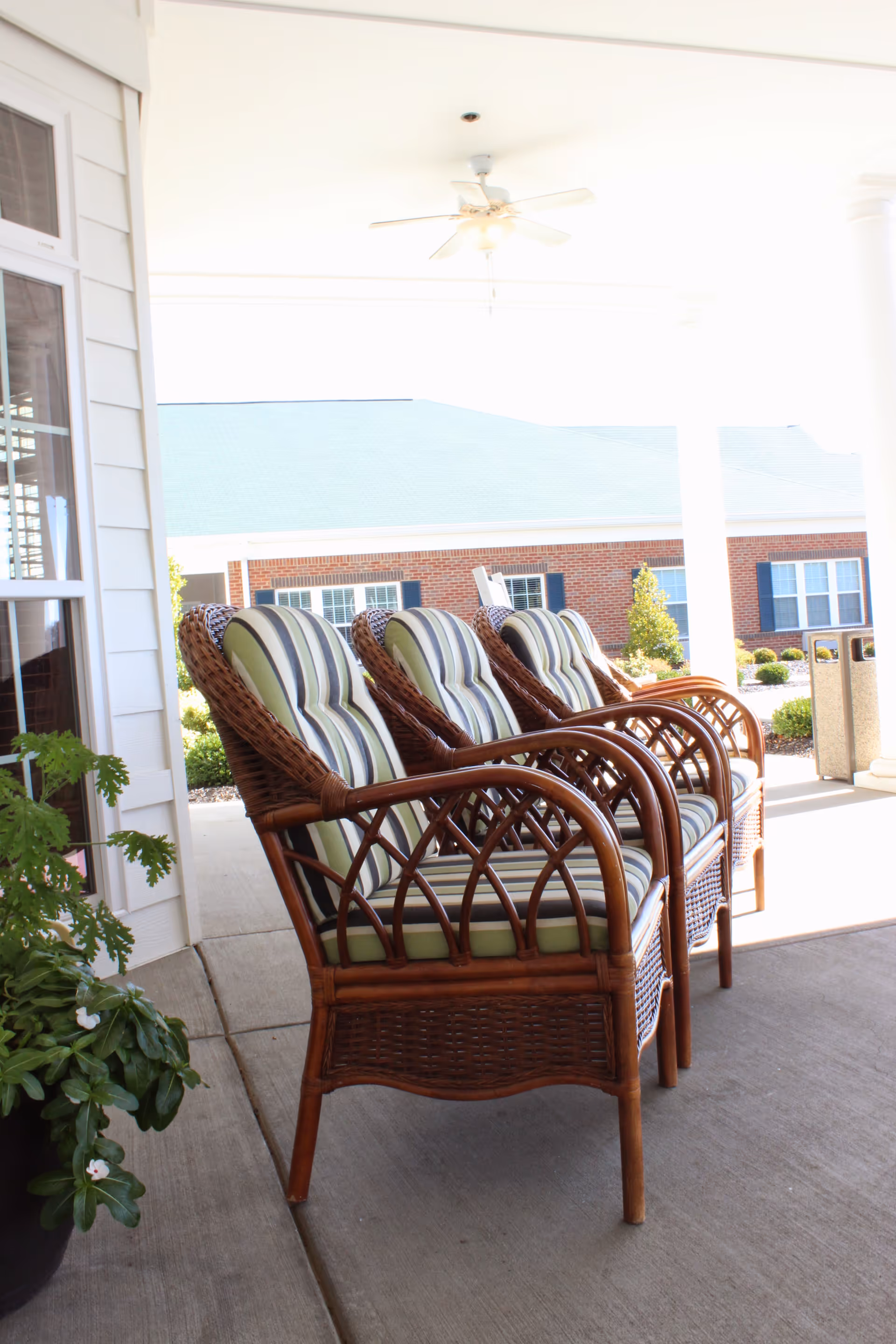 Three wicker chairs with striped cushions arranged on a covered porch with a ceiling fan above. In the background, there is a brick building with windows and some landscaping.