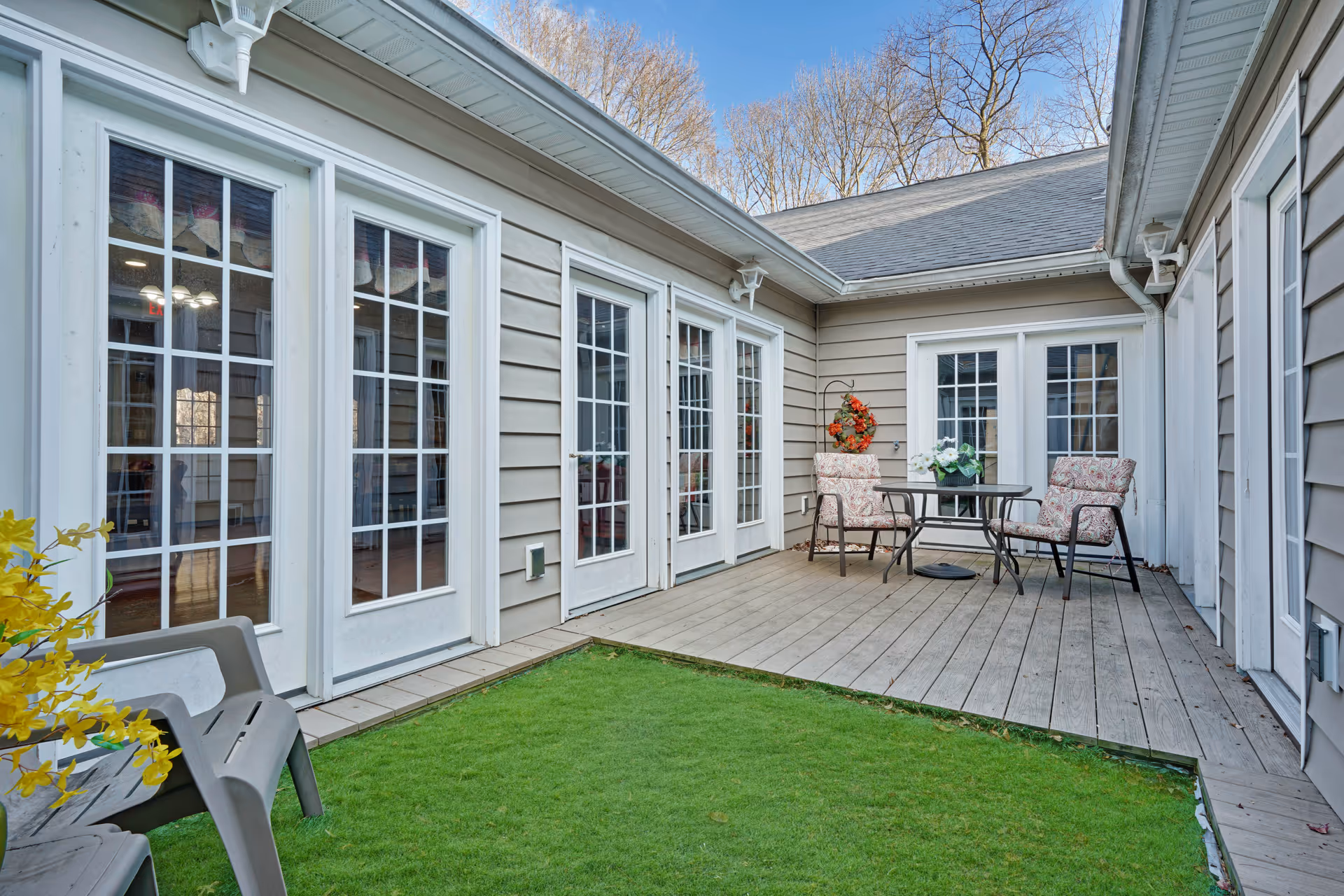 Outdoor patio area with wooden decking and artificial grass, surrounded by beige siding walls with multiple white-framed glass doors and windows. There is a small round table with two cushioned chairs and a flower arrangement on the table. Two plastic chairs and a potted plant with yellow flowers are also visible.