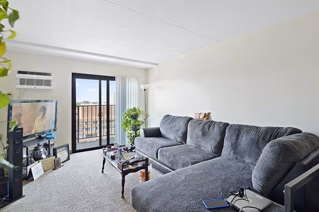 A living room with a large gray sectional sofa, a wooden coffee table with various items on it, a flat-screen TV mounted on the wall, a tall floor lamp, and a sliding glass door leading to a balcony with a view of nearby buildings.