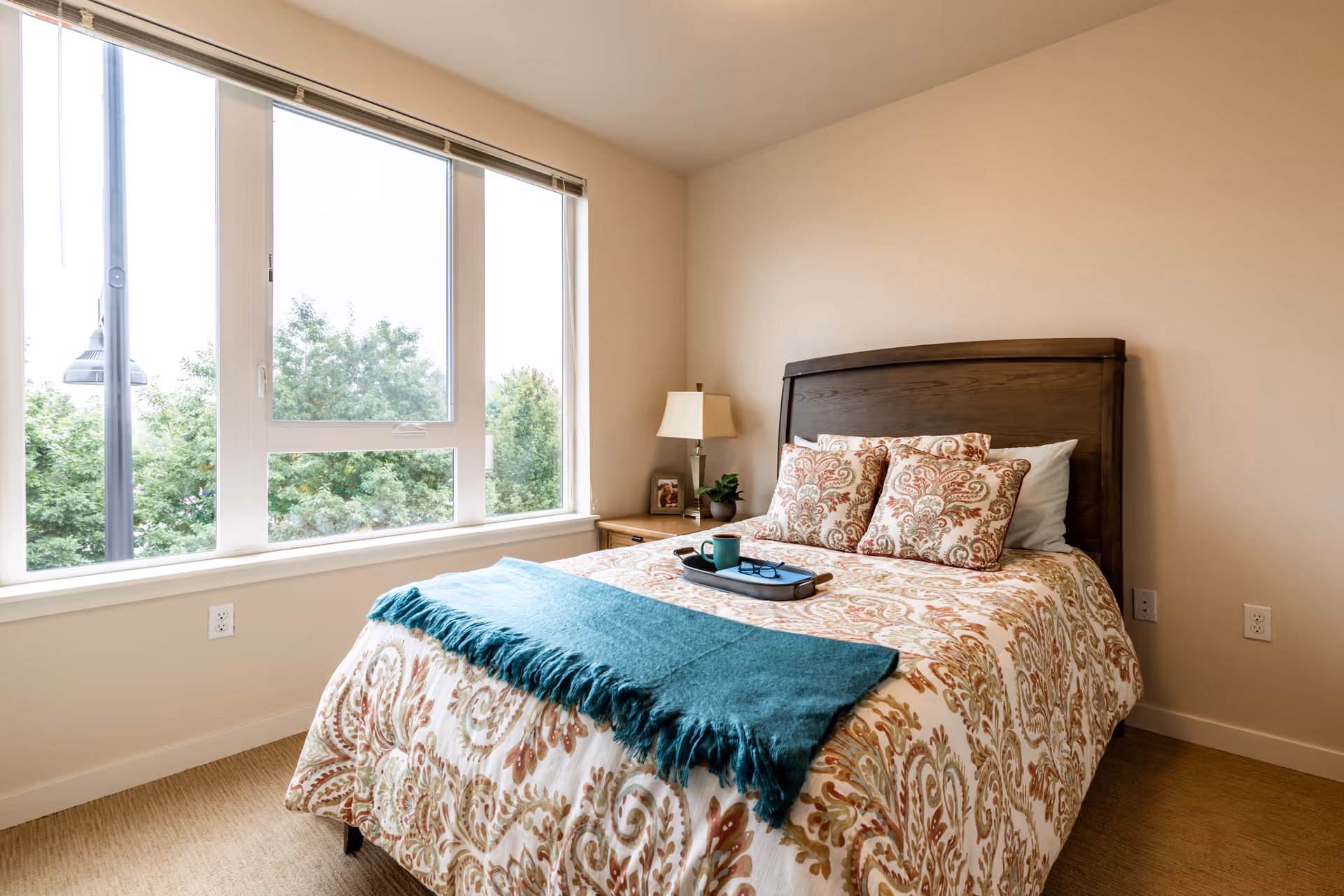 A cozy bedroom with a large window showing green trees outside. The bed has a wooden headboard and is covered with a patterned bedspread and matching pillows. A teal throw blanket is draped across the foot of the bed, with a tray holding a cup and glasses on top. A nightstand with a lamp, a small plant, and a framed photo is beside the bed.