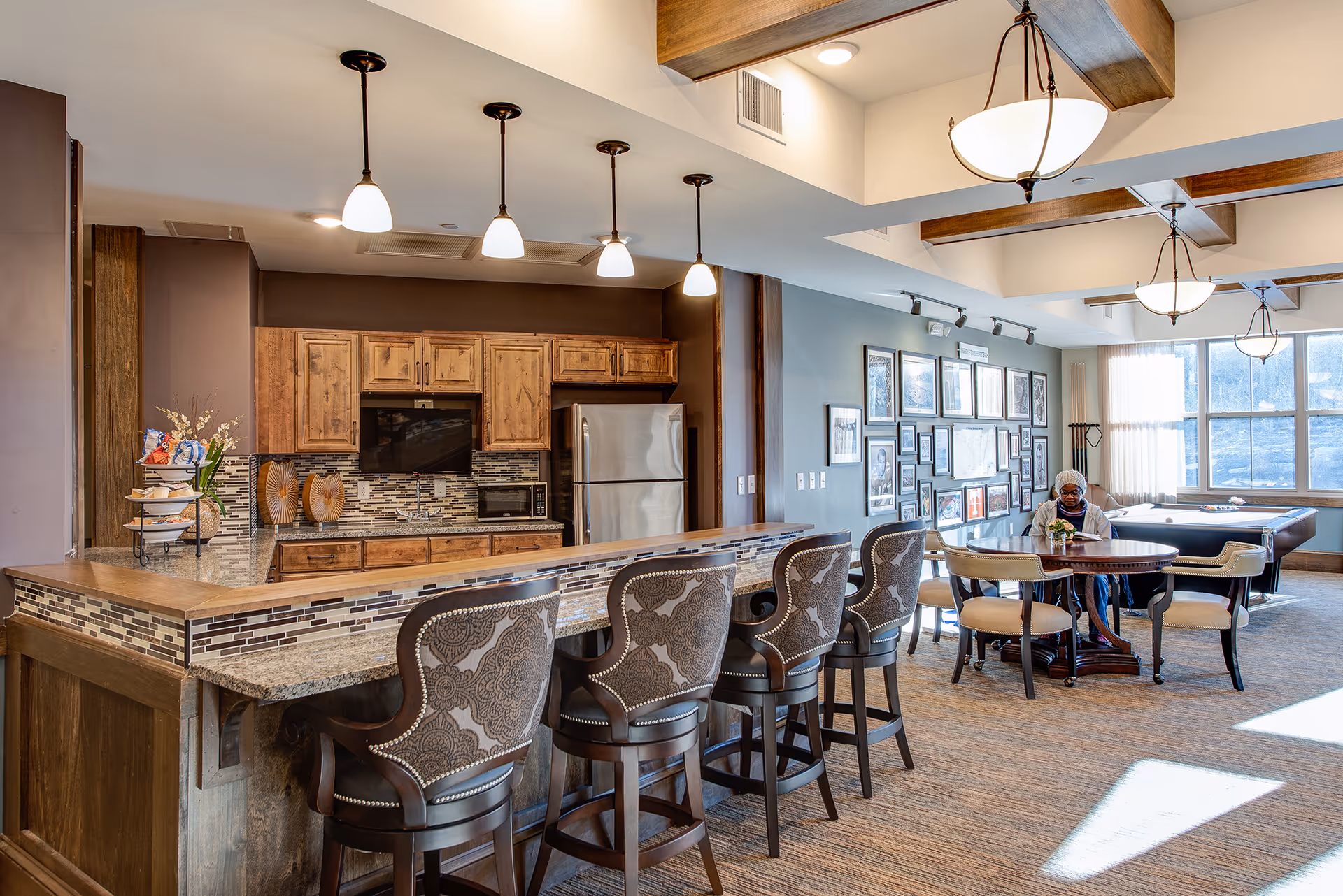 A spacious common area in a senior living facility featuring a kitchen with wooden cabinets, a granite countertop bar with five patterned high-back chairs, and pendant lights hanging from the ceiling. In the background, there is a seating area with a round table and chairs, a person sitting at the table, a wall decorated with framed pictures, and a pool table near large windows letting in natural light.