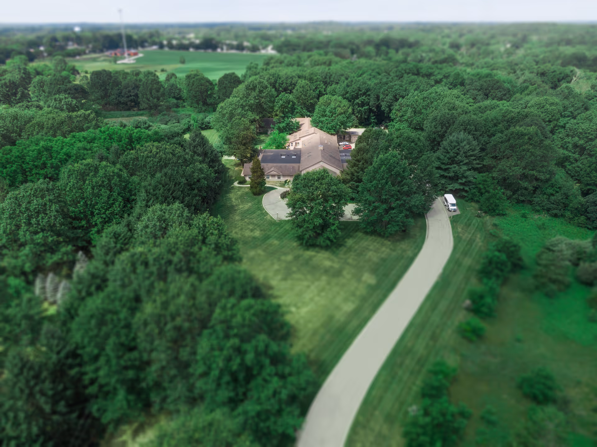 Aerial view of a single-story building surrounded by dense green trees and a well-maintained lawn. A curved driveway leads to the building, and a white vehicle is parked near the entrance. The area appears peaceful and secluded with a large expanse of greenery.
