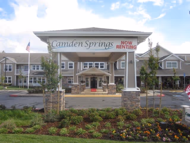 Entrance of Camden Springs senior living facility with a covered drop-off area displaying a sign that reads 'Camden Springs NOW RENTING'. The building is two stories with multiple windows and surrounded by landscaped greenery and flowers.