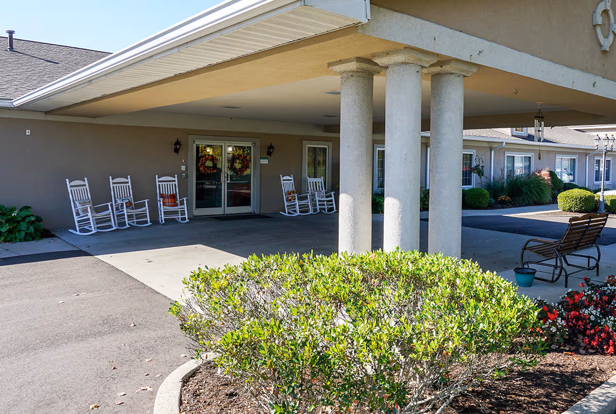 Entrance area of a senior living facility with a covered drop-off zone supported by three large columns. Several white rocking chairs are lined up against the building wall near double glass doors decorated with wreaths. There is a bench and some potted plants on the right side, and well-maintained bushes and flowers in the foreground.