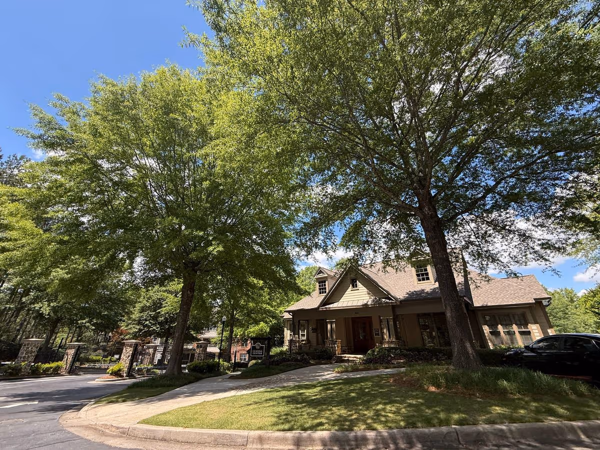 Front exterior of a clubhouse-style building with a driveway, large shade trees, and a manicured lawn under a blue sky.