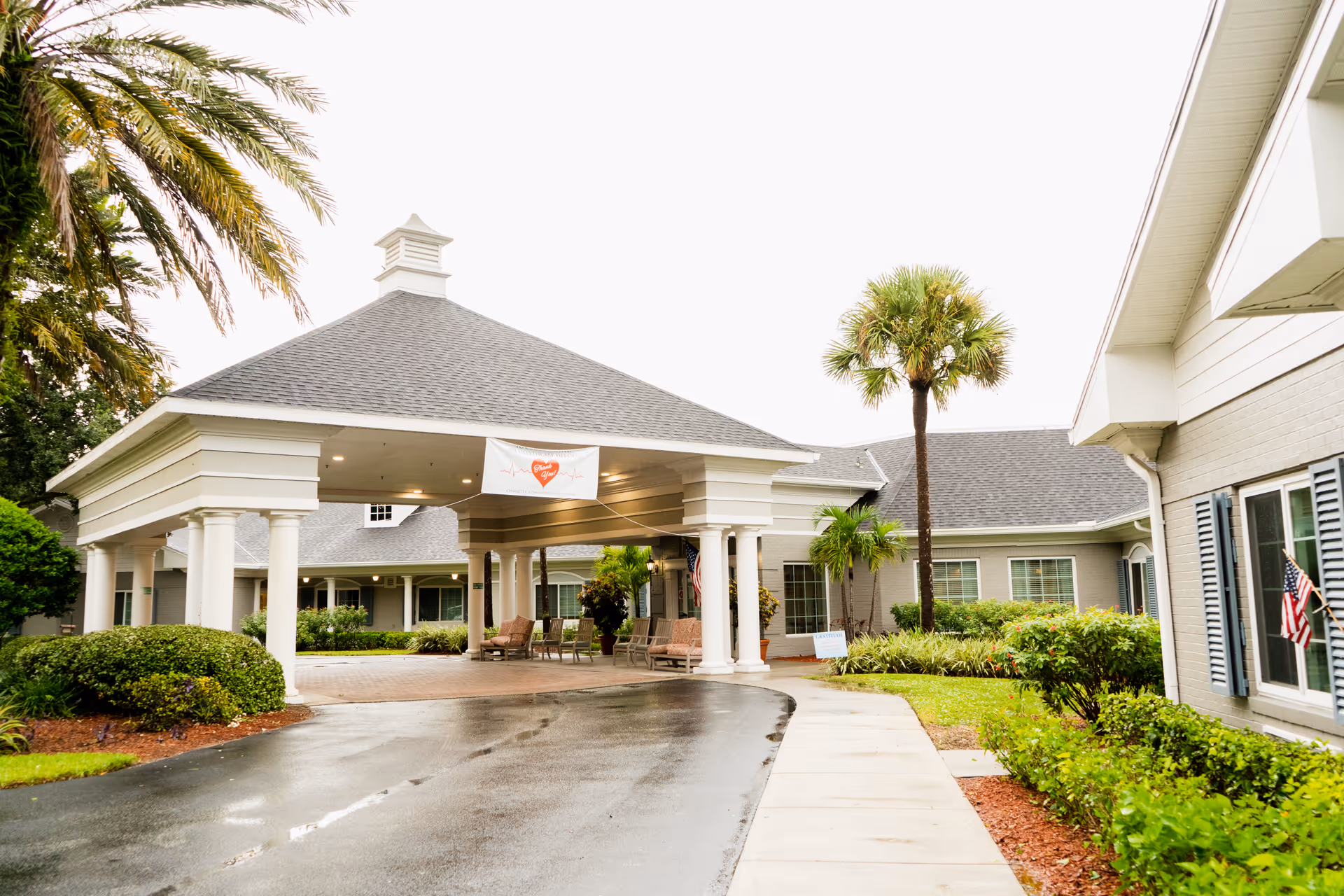 Entrance to a senior living facility with a covered drop-off area supported by white columns. The building is surrounded by well-maintained landscaping including palm trees and bushes. There is a banner hanging under the covered area and an American flag displayed near the windows.