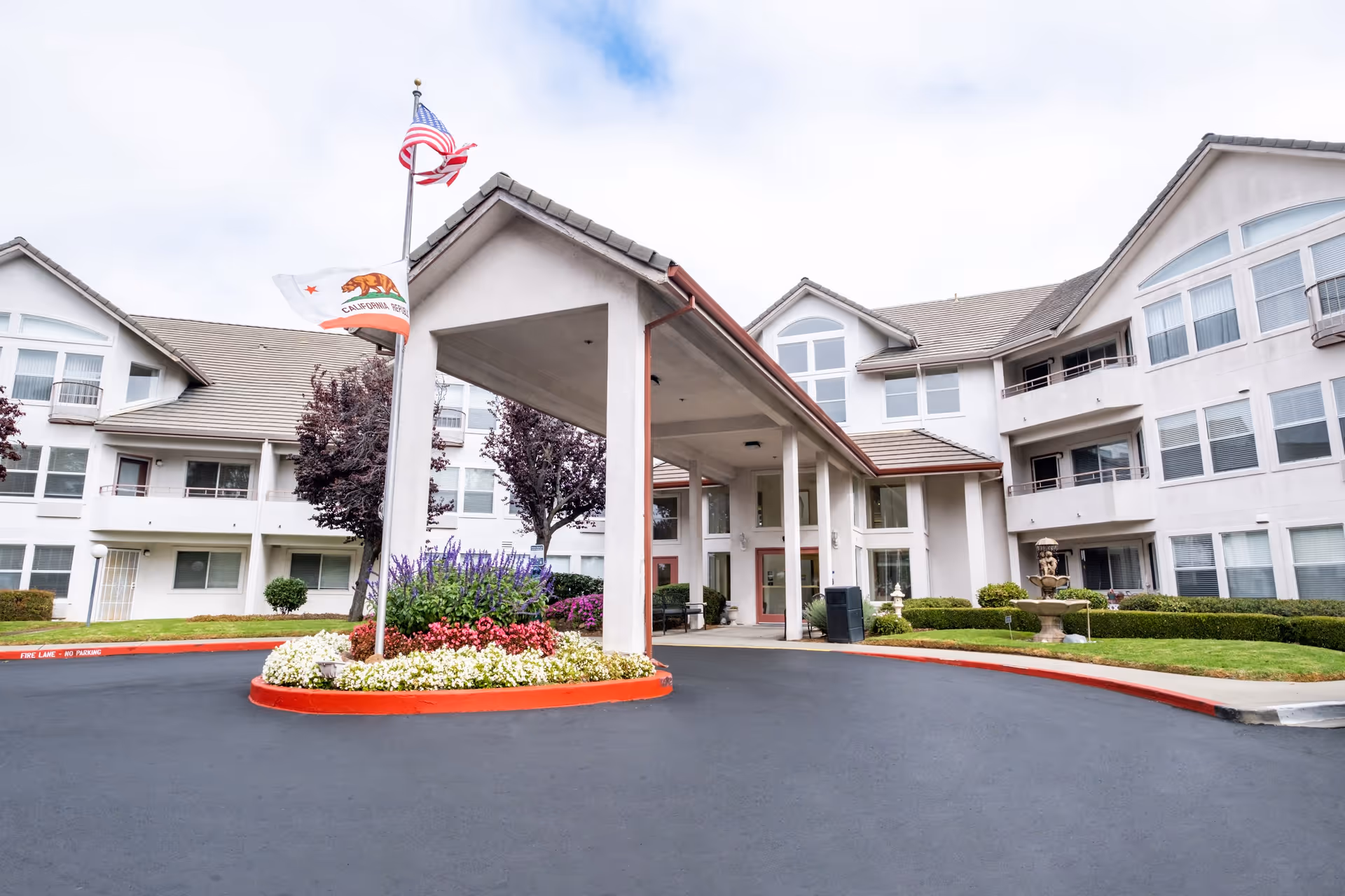 Exterior view of a senior living facility with a covered entrance, landscaped flower beds, and flags including the American and California state flags. The building has multiple windows and balconies with a fountain visible on the right side.