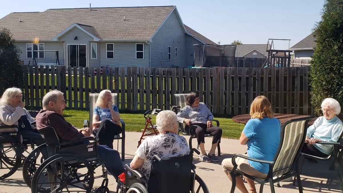 A group of elderly people, some in wheelchairs, sitting in a circle outdoors on a sunny day, engaging in conversation. They are seated on a paved area with a wooden fence and houses in the background.