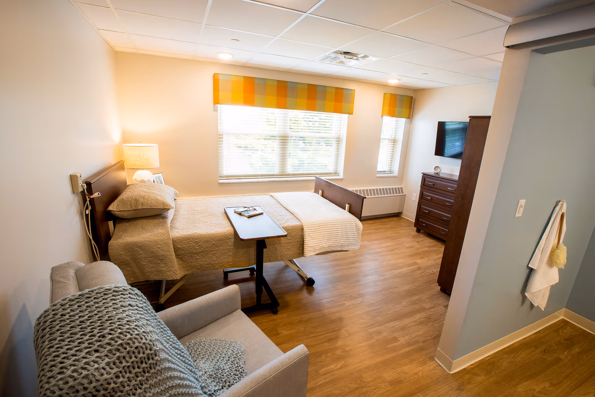 A bright and clean bedroom in a senior living facility with a single bed covered in beige bedding, a small overbed table with a book on it, a cushioned armchair with a knitted throw, a wooden dresser with a mounted flat-screen TV above it, and two windows with yellow and orange plaid valances letting in natural light.