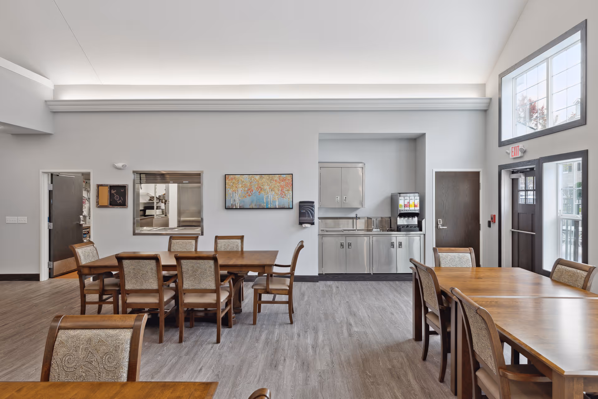 A bright dining room in a senior living facility with wooden tables and upholstered chairs. The room features large windows letting in natural light, a beverage dispenser on a stainless steel counter, and a colorful painting on the wall. There are two doors, one leading to another room and one exit door with an exit sign above it.