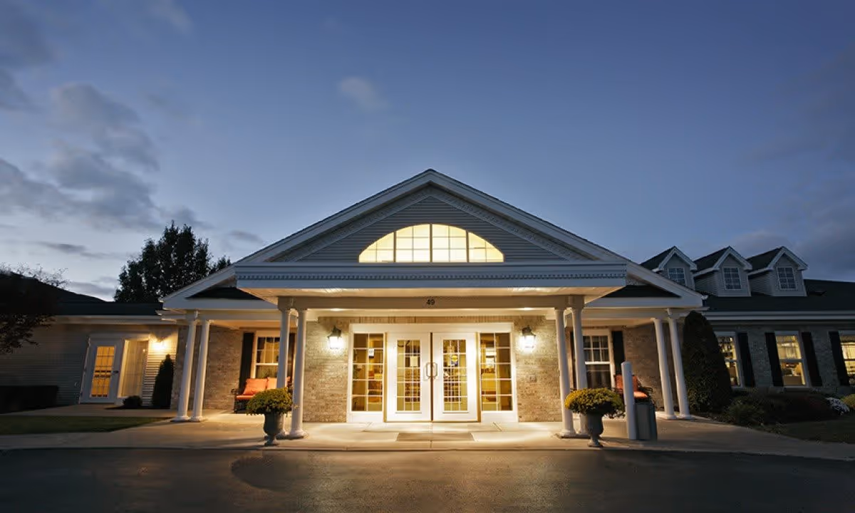 Front exterior view of a senior living facility named Tennyson Court at dusk, showing a well-lit entrance with double glass doors, white columns, and a triangular window above the entrance. There are potted plants on either side of the entrance and windows with lights on inside the building.