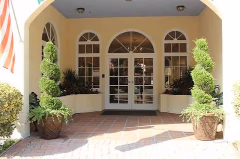 Exterior entrance of a retirement village with arched windows, glass doors, and potted topiary.