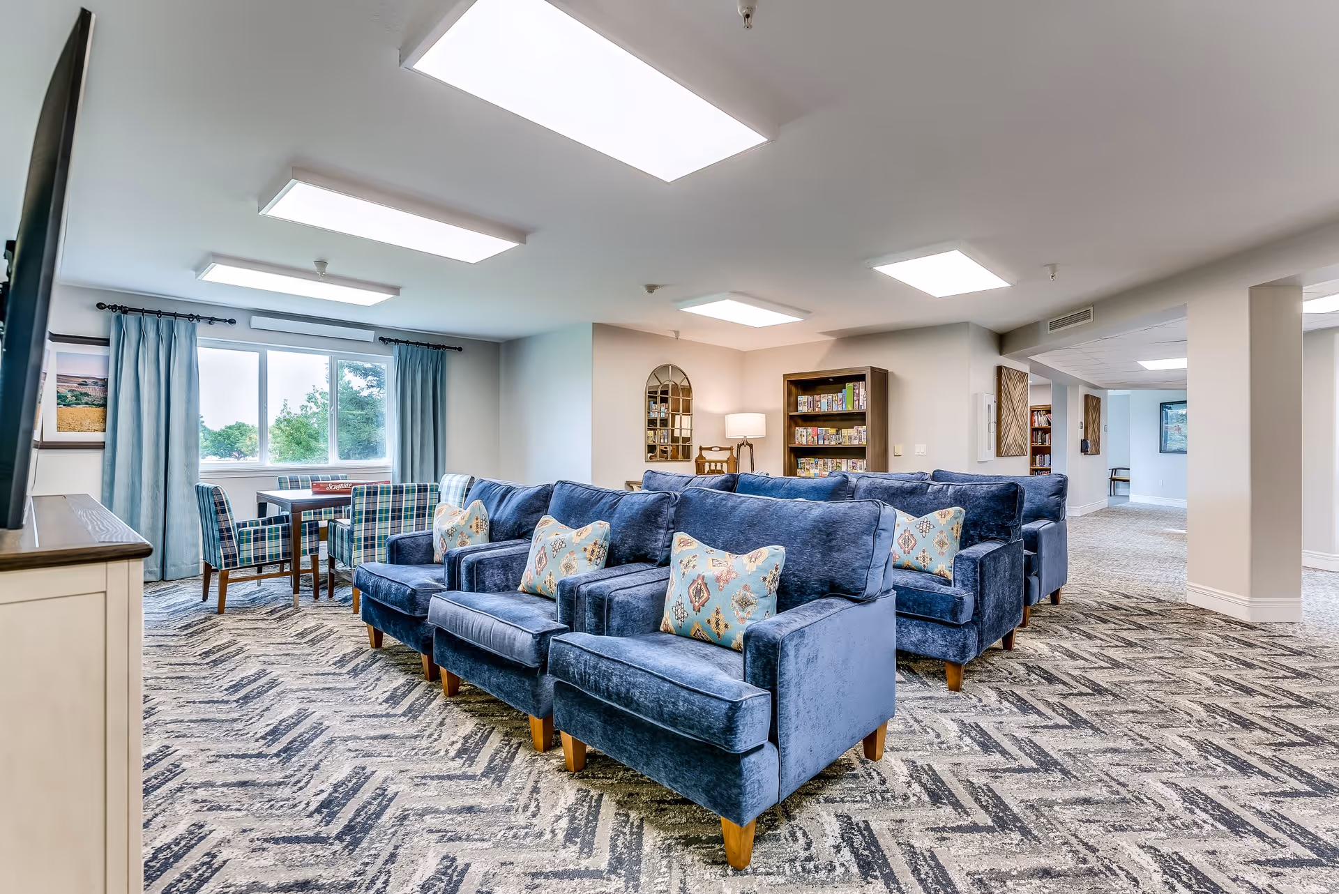 A bright and spacious senior living common area with multiple blue upholstered armchairs arranged in rows, each with a decorative pillow. There is a window with blue curtains letting in natural light, a small table with chairs near the window, a bookshelf filled with board games, and a lamp on a side table. The room has patterned carpet flooring and white walls with ceiling lights.