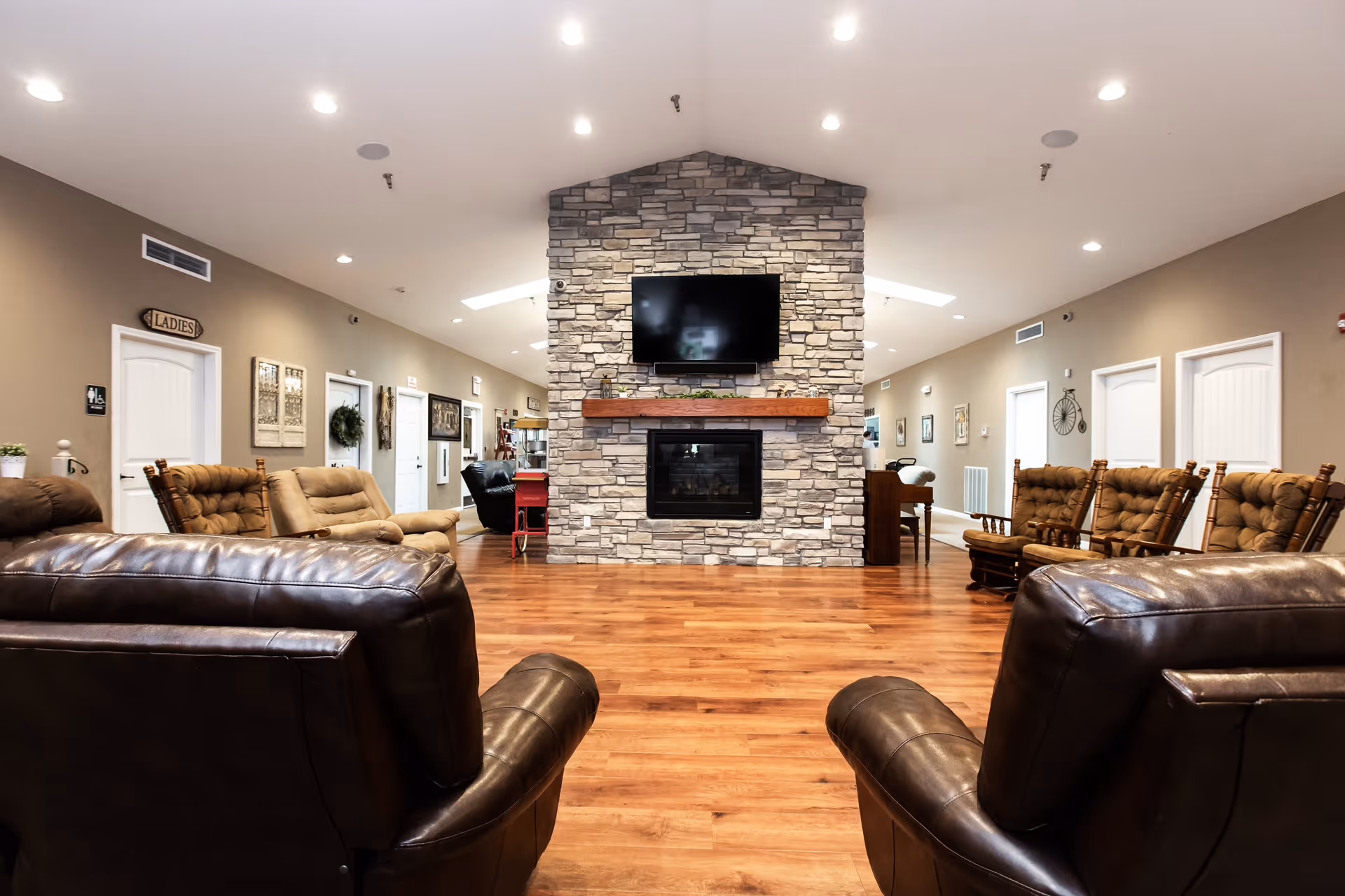 A spacious senior living facility common area with wooden flooring, multiple cushioned chairs and sofas arranged around a central stone fireplace with a mounted flat-screen TV above it. The walls are beige with white doors and various decorations, including a sign labeled 'LADIES' above one door.