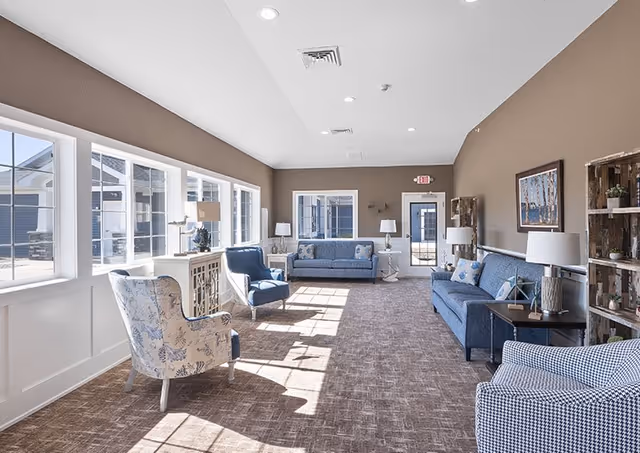 A bright and spacious living room in an assisted living facility with large windows letting in natural light. The room features multiple blue upholstered sofas and armchairs arranged along the walls, with side tables and lamps. The walls are painted brown with white trim, and there are decorative shelves and framed artwork on the walls. The carpet is a neutral brown tone, and the ceiling is vaulted with recessed lighting.