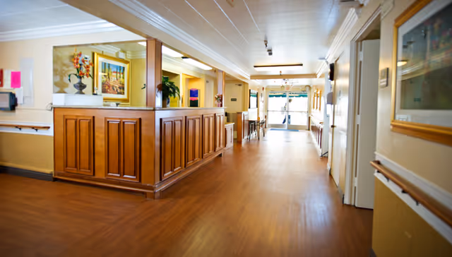 A well-lit nursing center hallway with wooden flooring and a wooden reception desk on the left side. The walls are painted in light colors with handrails along both sides. There are framed pictures and plants decorating the space, and glass doors are visible at the far end of the hallway.