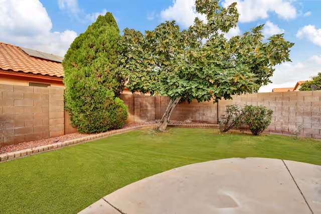A small, enclosed backyard with a green lawn, a concrete patio area, a tree with broad leaves, a tall bush, and a brown brick wall surrounding the yard under a partly cloudy blue sky.