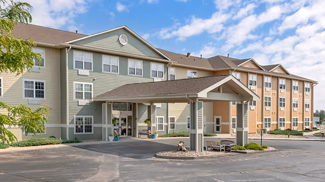 Exterior view of a three-story senior living facility building with a covered entrance driveway. The building has multiple windows and a combination of beige and light green siding. There are some small landscaped areas with bushes and a tree near the entrance. The sky is partly cloudy.