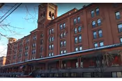 Red brick multi-story historic apartment building with a tower, arched windows, and a covered porch with snow along the sidewalk.