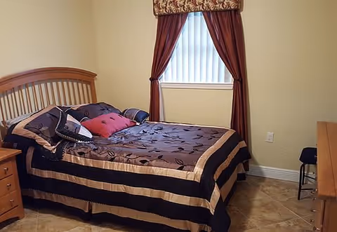 Small bedroom with a double bed dressed in patterned bedding, a wooden headboard, nightstand and dresser beneath a curtained window.