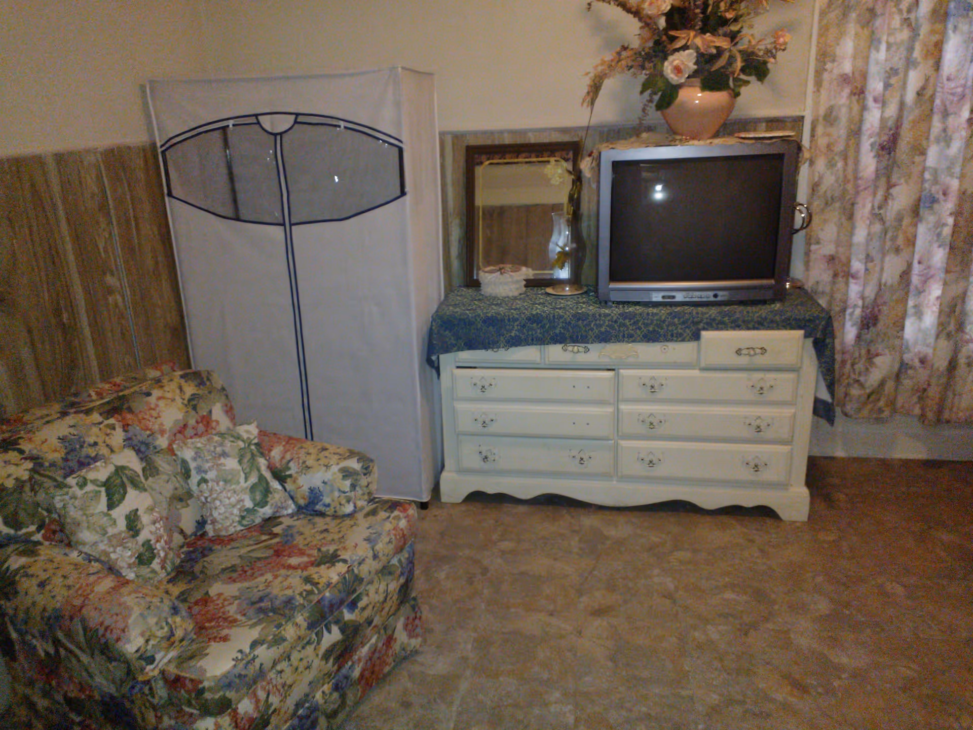 A cozy room with a floral-patterned armchair, a white dresser covered with a blue cloth, an old-style CRT television on top, a mirror, a glass lamp, and a flower vase. There is a fabric wardrobe or storage unit next to the dresser, and floral curtains hang on the right side.