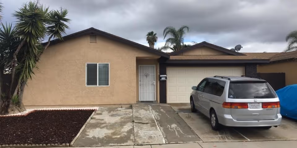 Single-story stucco house front with a garage, driveway and a silver minivan parked under a cloudy sky.