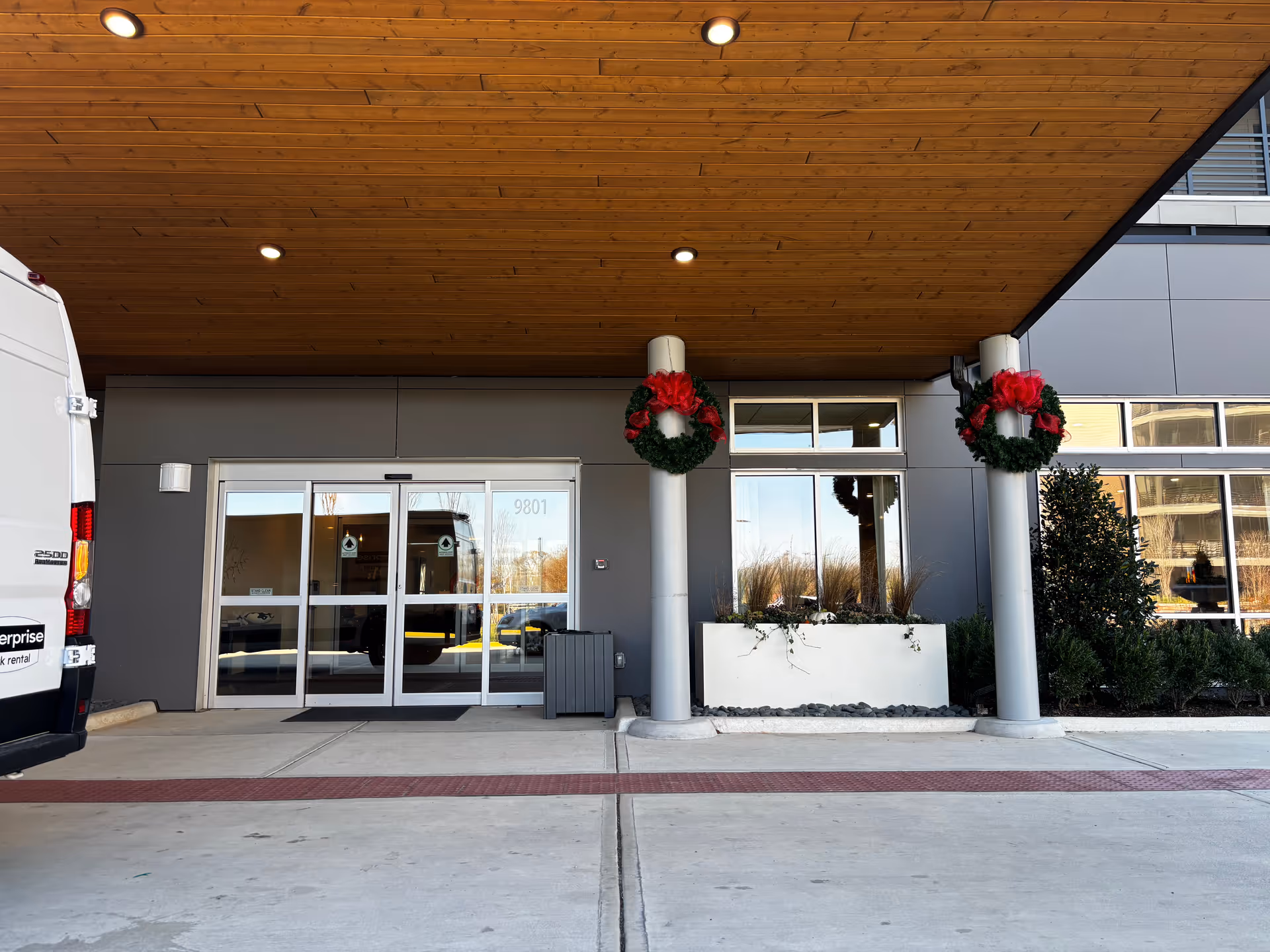Covered main entrance with sliding glass doors under a wooden canopy, holiday wreaths on columns, and a van parked at the curb.
