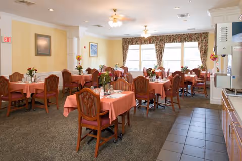 A dining room with multiple tables covered in pink tablecloths, each set with chairs and small floral centerpieces. The room has large windows with floral curtains allowing natural light to fill the space. Ceiling fans and framed artwork are visible on the walls.