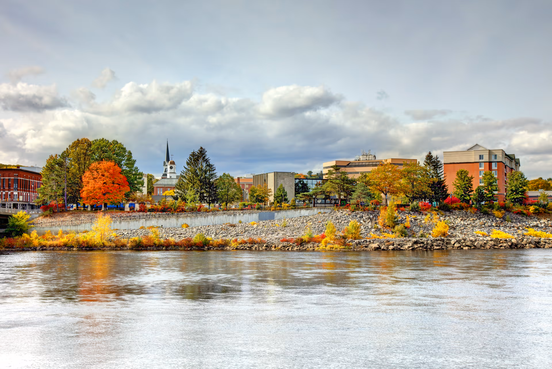 View of a riverside area with autumn-colored trees and a mix of buildings including a church with a steeple and multi-story structures under a cloudy sky.