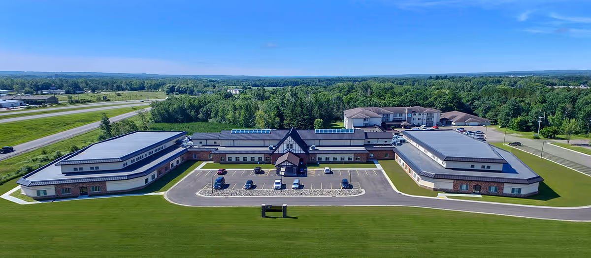 Aerial view of Little Falls Health Services facility showing a large, modern building with multiple wings surrounded by green lawns and parking areas, set against a backdrop of trees and a clear blue sky.
