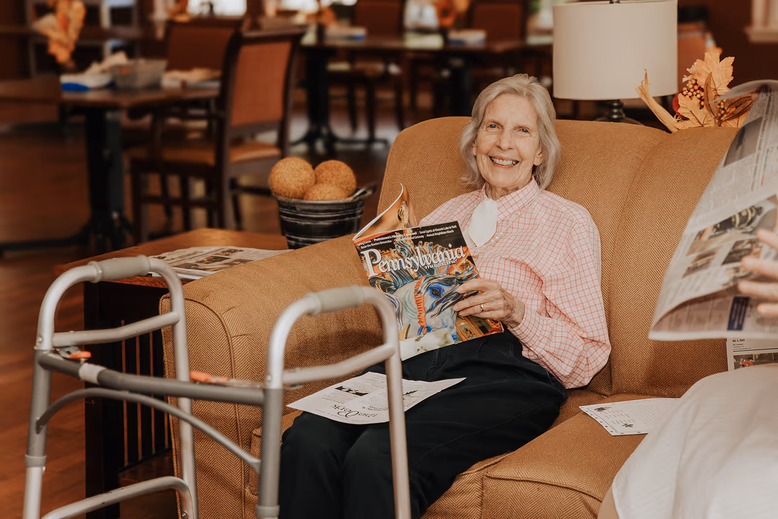 An elderly woman sits on a couch in a communal lounge smiling while reading a magazine, with a walker in the foreground.