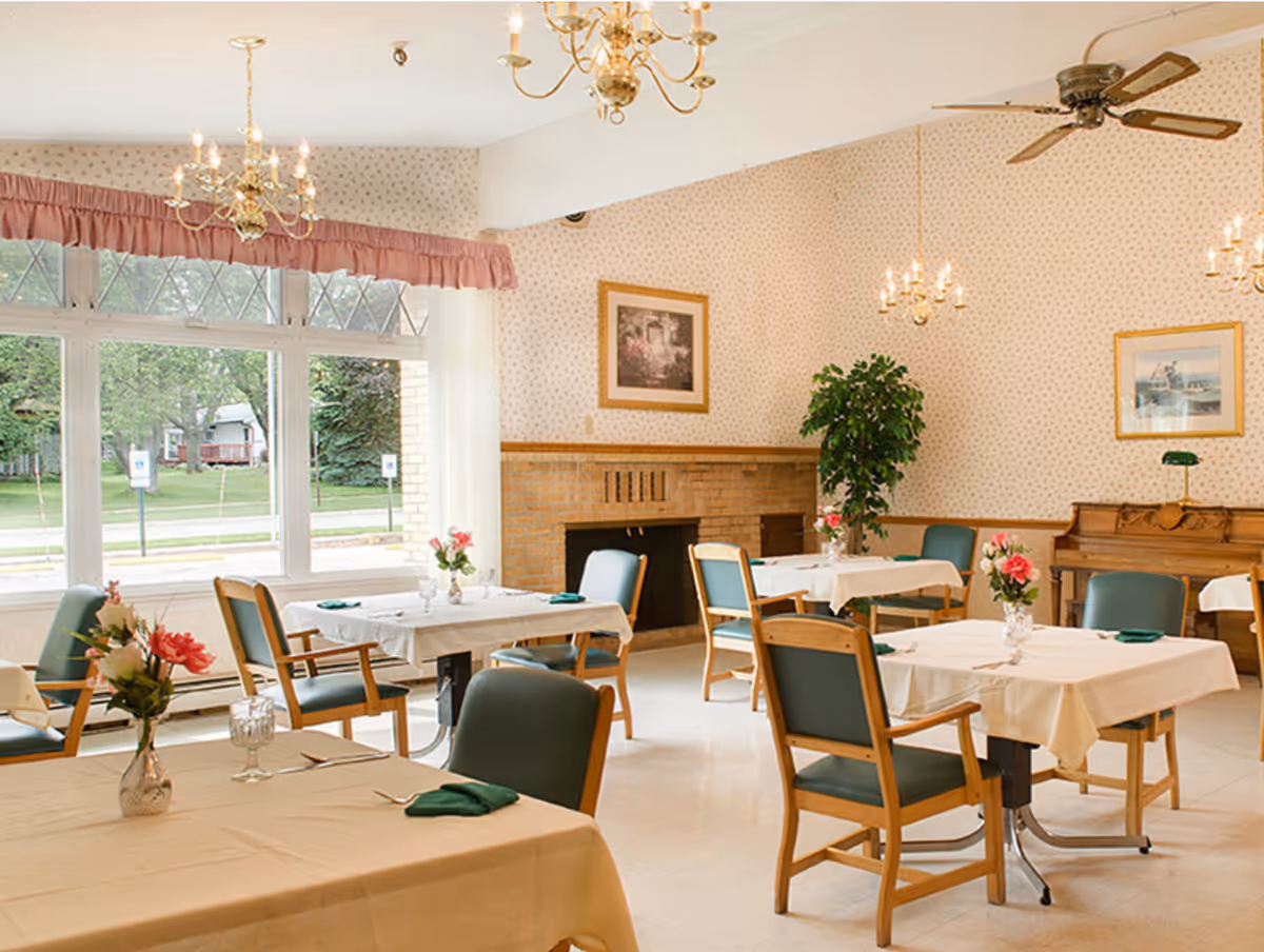 A bright dining room with several tables covered in white tablecloths, each set with green napkins and floral centerpieces. The room features large windows with pink valances, chandeliers, a ceiling fan, framed artwork on the walls, a fireplace, and a piano in the corner.