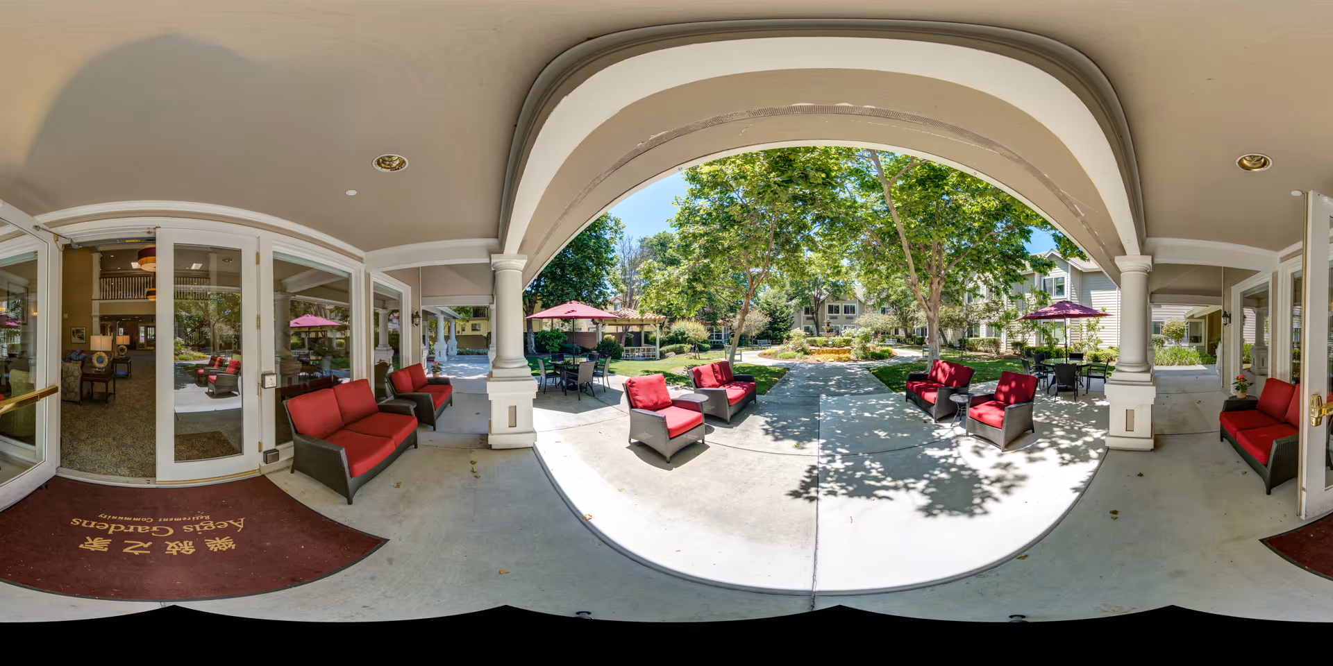 Covered outdoor patio area with red cushioned chairs and tables with umbrellas, overlooking a garden with trees and a walking path. The entrance to a building with glass doors and windows is visible on the left side, with a maroon mat that reads 'Aegis Gardens'.