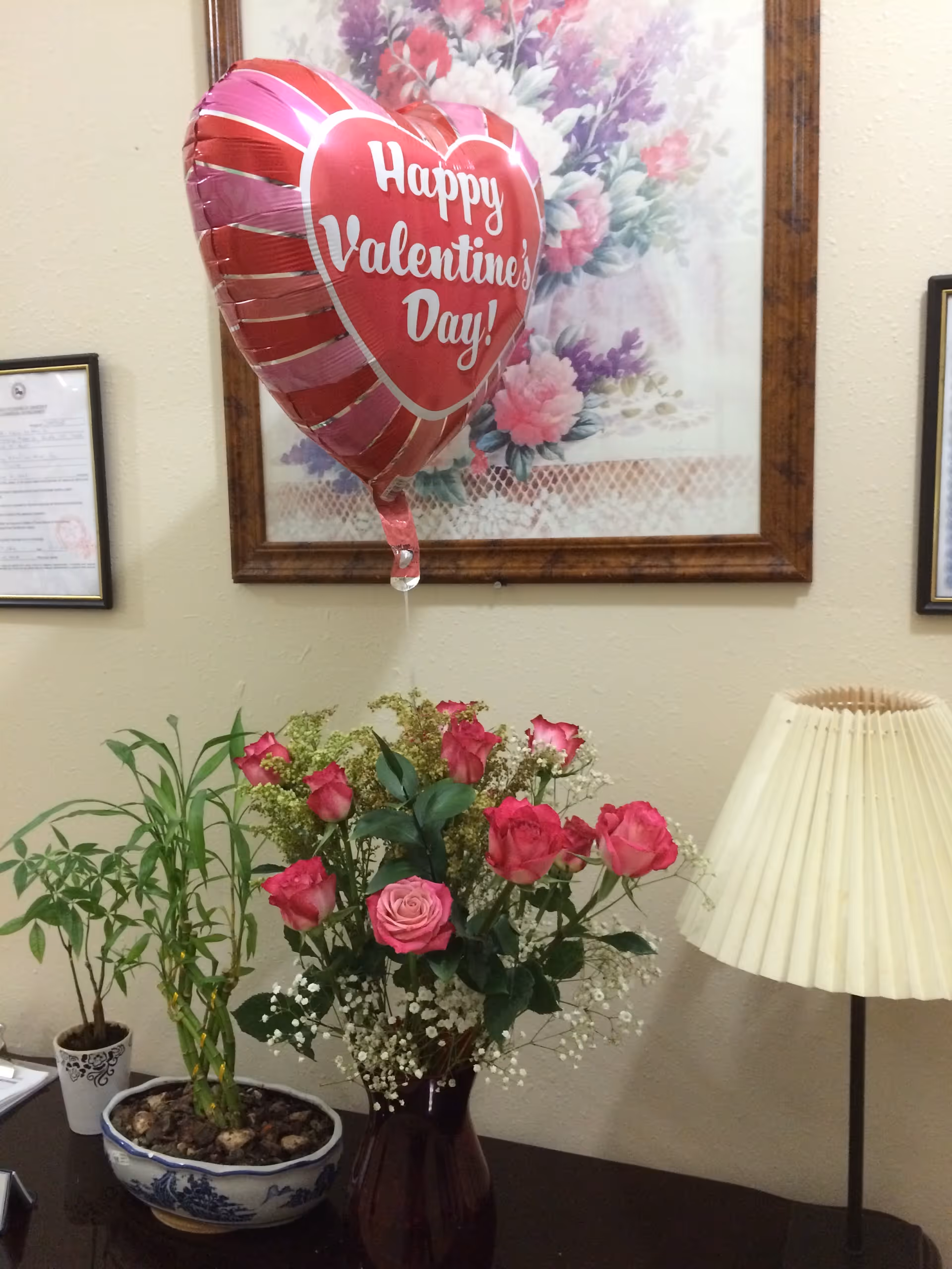 A heart-shaped "Happy Valentine's Day!" balloon floats above a vase of pink roses, potted plants, and a lamp on a table under a framed floral painting.