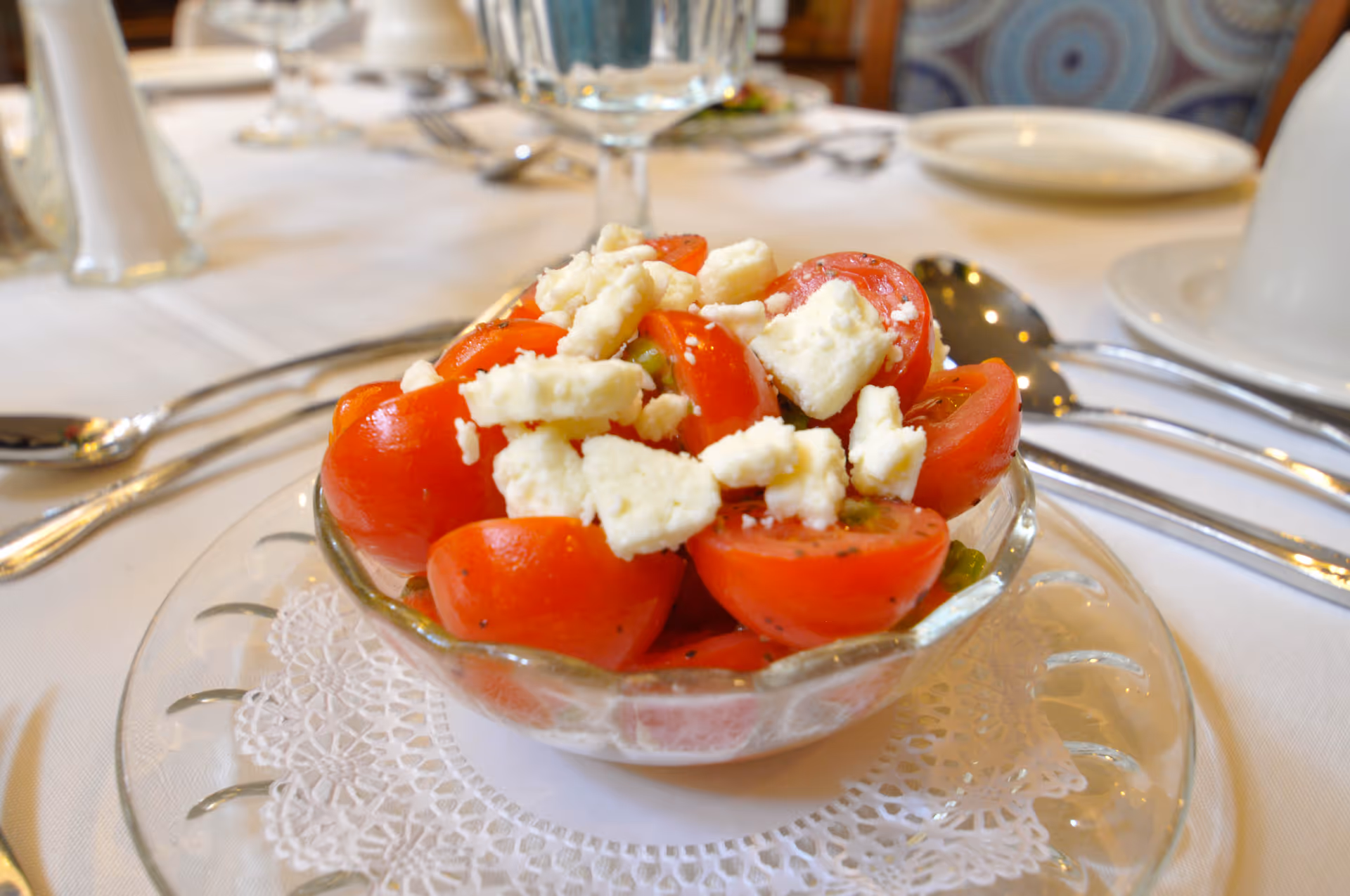 Bowl of halved cherry tomatoes topped with crumbled cheese on a place setting at a dining table.