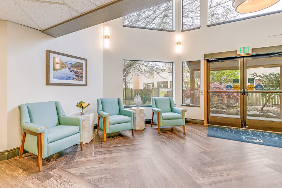 Bright seating area in a senior living lobby with three mint-green armchairs, side tables, a lamp, and glass entry doors.