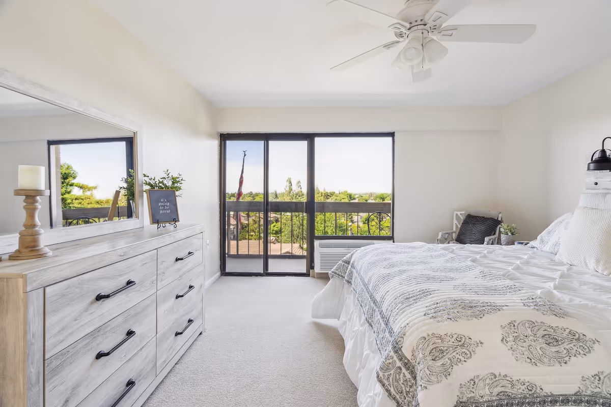 Bright bedroom with a bed, patterned bedding, dresser and ceiling fan opening onto a balcony through sliding glass doors.