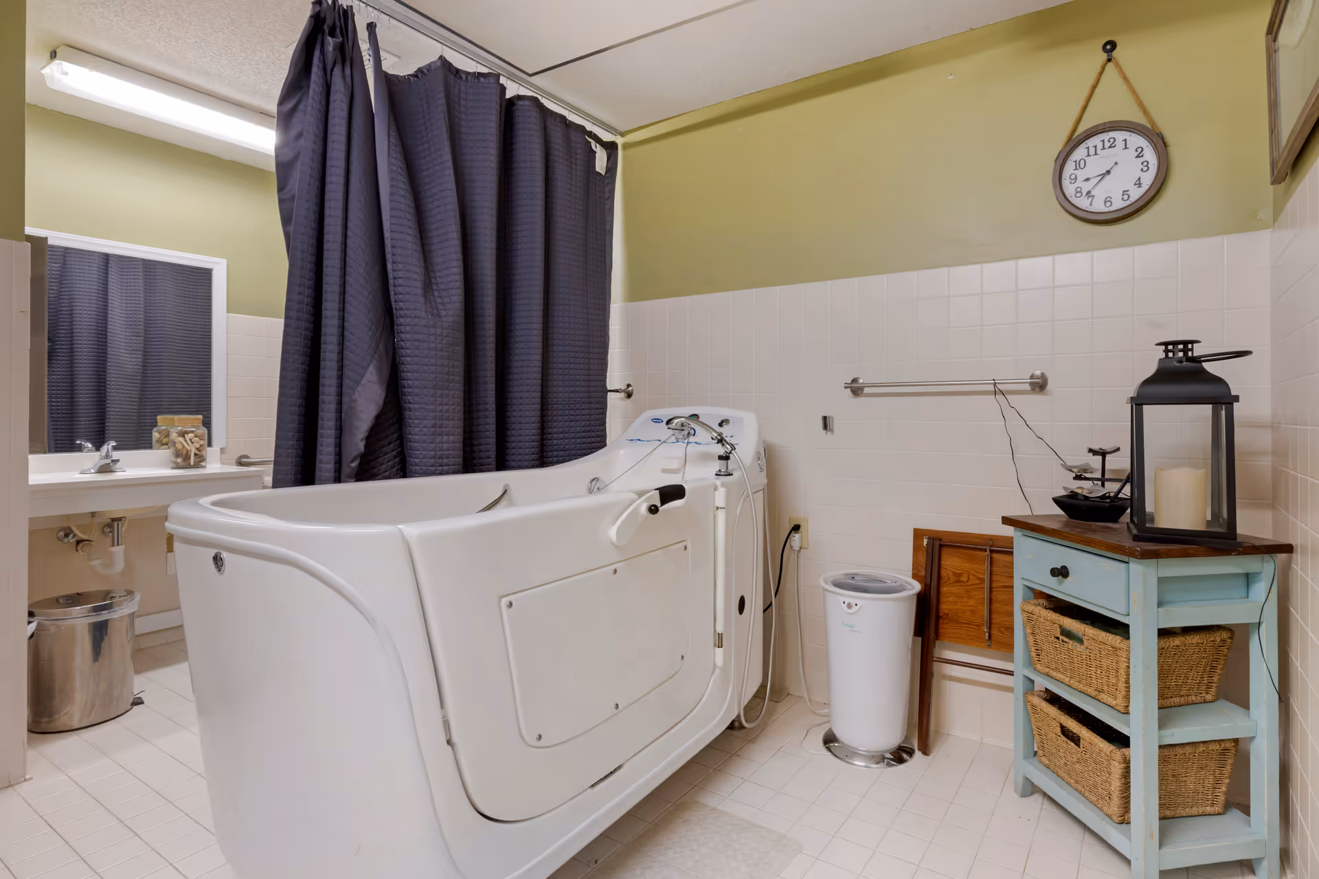 A bathroom featuring a walk-in bathtub with a door and handrails, a dark shower curtain, a sink with a mirror, a small blue wooden cabinet with wicker baskets, a black lantern with a candle on top, a wall clock, and a trash can.