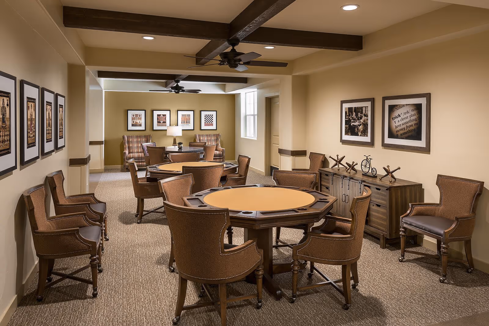 A cozy interior room with two octagonal card tables surrounded by brown upholstered chairs. The room features beige walls adorned with framed artwork, a wooden sideboard with decorative items, ceiling fans with wooden beams, and a carpeted floor. In the background, there are two armchairs and a small table with a lamp near a window.