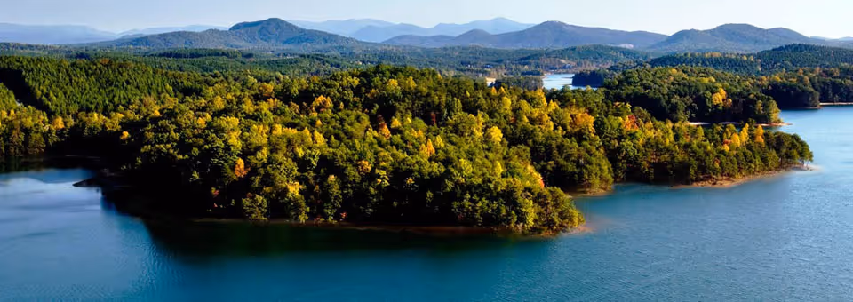 A scenic aerial view of a forested peninsula surrounded by blue water with distant mountain ranges under a clear sky.