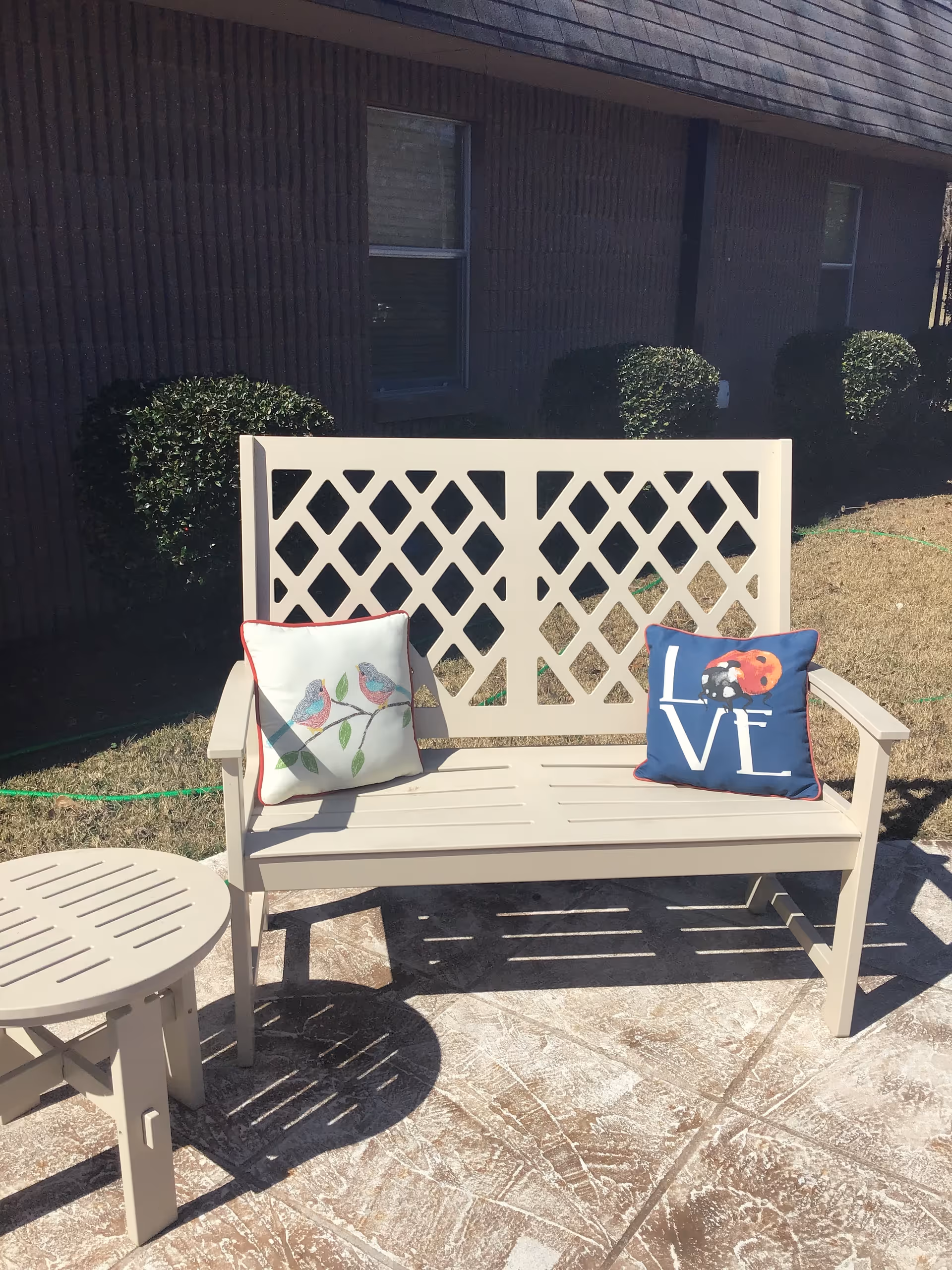 Beige outdoor bench with a lattice back and two decorative pillows on a sunlit patio in front of a brick building.