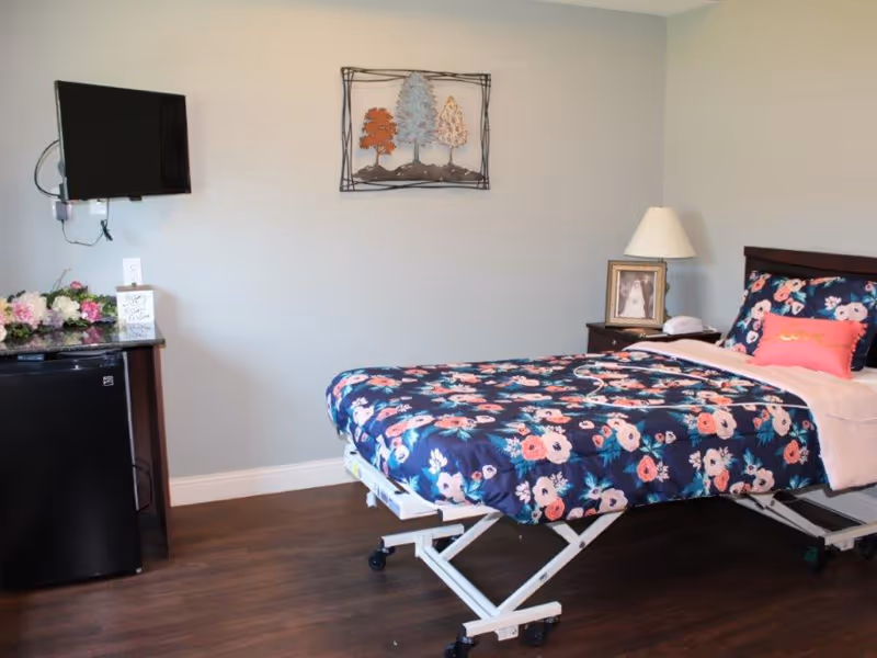 A bedroom in a care facility featuring a hospital-style bed with a floral comforter and a pink pillow with the word 'love'. The room has a wooden floor, a small black refrigerator topped with flowers and a card, a wall-mounted flat-screen TV, a nightstand with a lamp and a framed photo, and a decorative metal wall art piece depicting three trees.