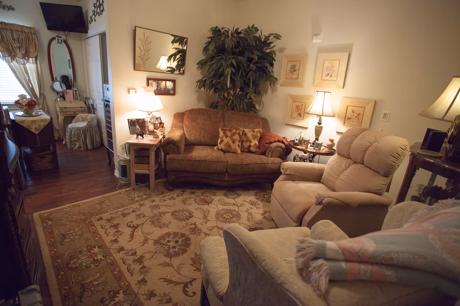 Cozy living room with a patterned brown loveseat, a beige recliner, and an armchair with a folded blanket. The room features a large floral area rug, two table lamps, framed floral artwork on the wall, a large potted plant, and a mirror above a small side table with family photos. In the background, there is a small dining area with a table, chairs, and a vanity with a mirror and stool near a window with draped curtains.