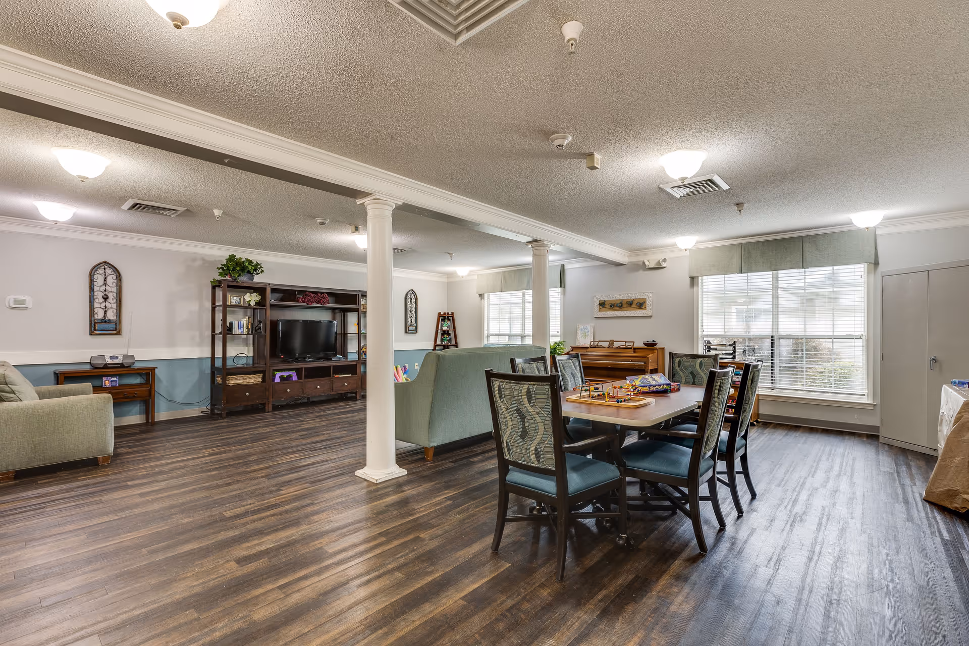 A spacious common living area with wooden flooring, featuring a dining table with six chairs, two green sofas, a wooden entertainment center with a TV, and large windows allowing natural light to fill the room. The room has white walls with blue wainscoting and decorative wall hangings.