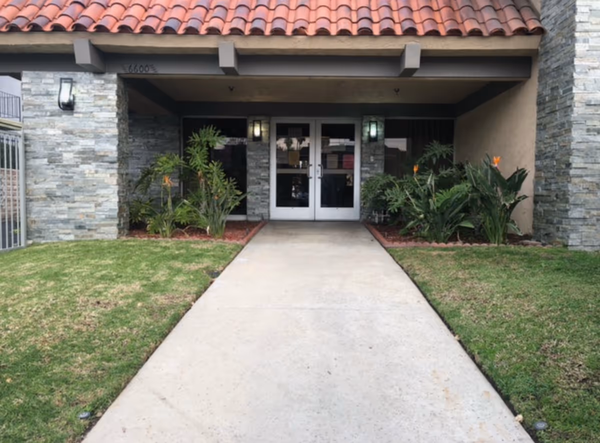 Concrete walkway leading to double glass entrance doors of a stone-clad building with landscaping and a tiled overhang.