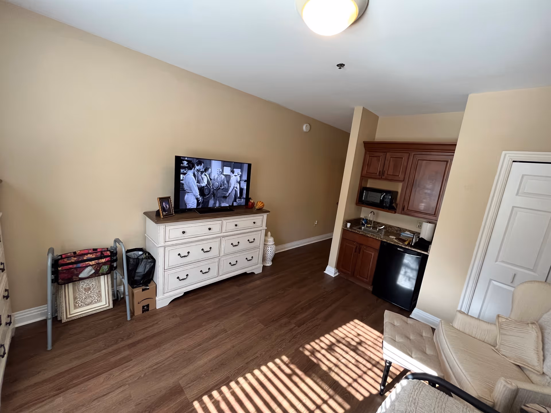 A cozy living area with a white dresser holding a flat-screen TV displaying a black and white movie. Next to the dresser is a walker and some framed artwork. The room has wooden flooring and beige walls. To the right, there is a small kitchenette with wooden cabinets, a microwave, a sink, and a mini refrigerator. A beige armchair with cushions is partially visible in the foreground.