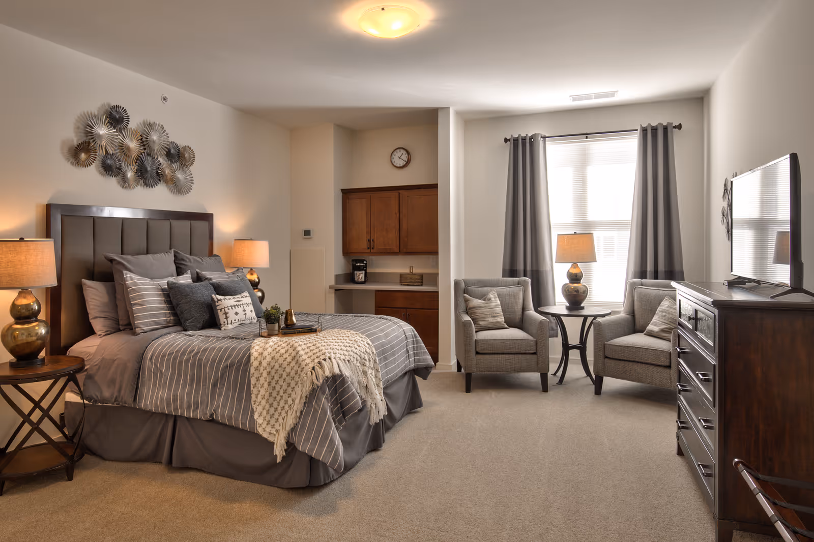 A cozy bedroom in a senior living facility with a large bed dressed in gray and striped bedding, multiple pillows, and a throw blanket. Two bedside tables with matching lamps flank the bed. On the wall above the bed is a decorative metal wall art piece. To the right, there are two gray armchairs with pillows and a small round table with a lamp between them, positioned in front of a window with gray curtains. A dark wooden dresser with a flat-screen TV on top is on the right side of the room. The room has beige carpet and neutral-colored walls, with a small kitchenette area featuring wooden cabinets and a coffee maker in the background.