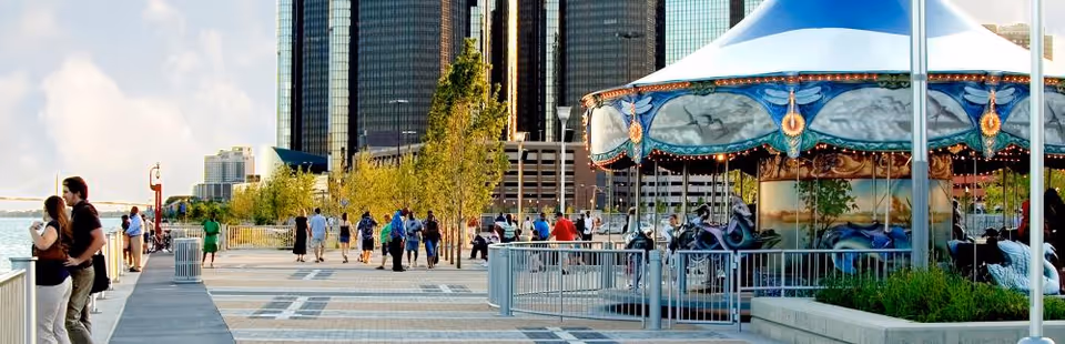 A waterfront promenade with people walking and standing near railings. There is a carousel with blue and white canopy on the right side. Tall buildings and trees are visible in the background under a partly cloudy sky.